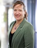 Porträt einer Person im grünen Sakko mit Halskette vor hellem, unscharfem Hintergrund.